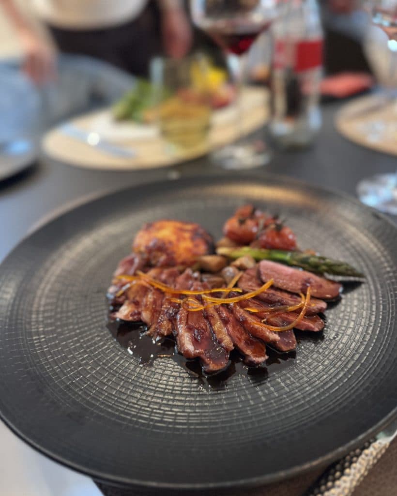 Op een zwart bord liggen plakjes eendenborst, overgoten met een donkere saus en gegarneerd met sinaasappelschil, vergezeld van gegrilde asperges, geroosterde tomaten, olijven en een goudbruine aardappelkoek – een verfijnde smaakbeleving uit het kanton Vaud, vlak bij het rustige meer van Morges. Op de achtergrond staat rode wijn klaar.