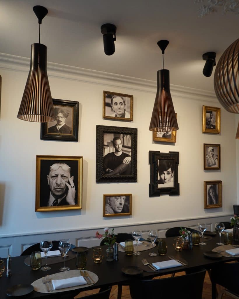 Een eetkamer in Morges met een lange zwarte tafel, gedekt met glazen, borden en servetten. Aan de muur erachter hangen ingelijste zwart-witportretten van beroemdheden. Boven dit elegante Vaud-tafereel hangen drie moderne hanglampen aan het plafond.