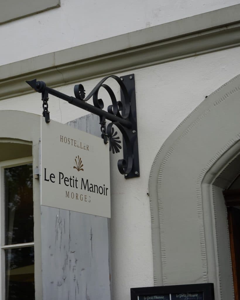Op een hangend bord staat "Le Petit Manoir Morges Hostellerie" te lezen, met een decoratieve metalen beugel, in het charmante Morges in het kanton Vaud. Het bord is bevestigd aan een wit gebouw met gewelfde raamkozijnen en een houten deur die onderaan gedeeltelijk zichtbaar is.