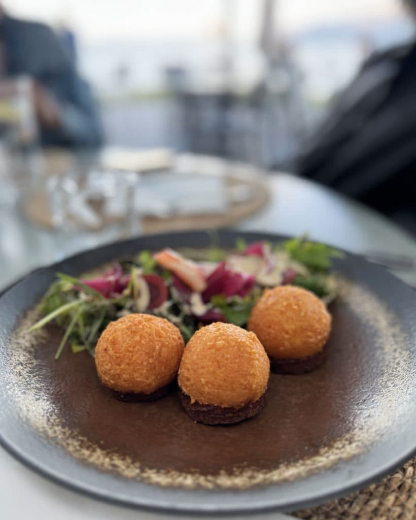 Drie goudkleurige, ronde gefrituurde balletjes liggen op een rij op een donker bord, vergezeld van een frisse salade met gemengde bladgroenten en rode groenten. De zacht vervaagde achtergrond roept het beeld op van een maaltijd aan het rustige meer van Morges in Vaud.