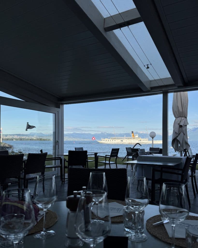 Een restaurant in Vaud met lege tafels en stoelen kijkt uit over het blauwe meer. Op de tafels staan glazen en liggen placemats, terwijl een witte veerboot met een Zwitserse vlag langs Morges vaart, omringd door bergen onder een gedeeltelijk bewolkte hemel.