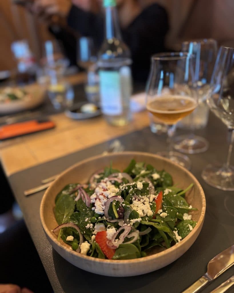 Op een restauranttafel in Morges, in het kanton Vaud, staat een houten schaal met verse salade met spinazie, fetakaas, rode uien en plakjes tomaat. Op de tafel staan glazen, een biertje, bestek en een glas water; op de achtergrond zijn mensen te zien die aan het eten zijn, maar die enigszins onscherp zijn.