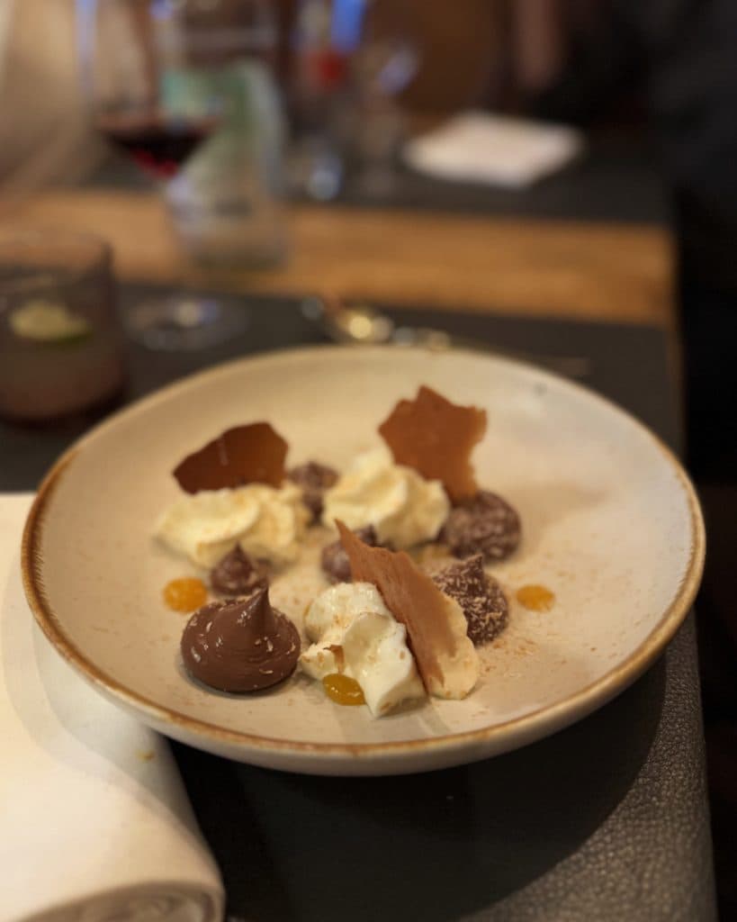 Een beige keramisch bord met toefjes slagroom, chocolademousse en sinaasappelsaus, gegarneerd met dunne stukjes karamel. Op de wazige achtergrond zijn een servet en drankjes te zien op een houten tafel in Morges, in het kanton Vaud, vlak bij het meer.