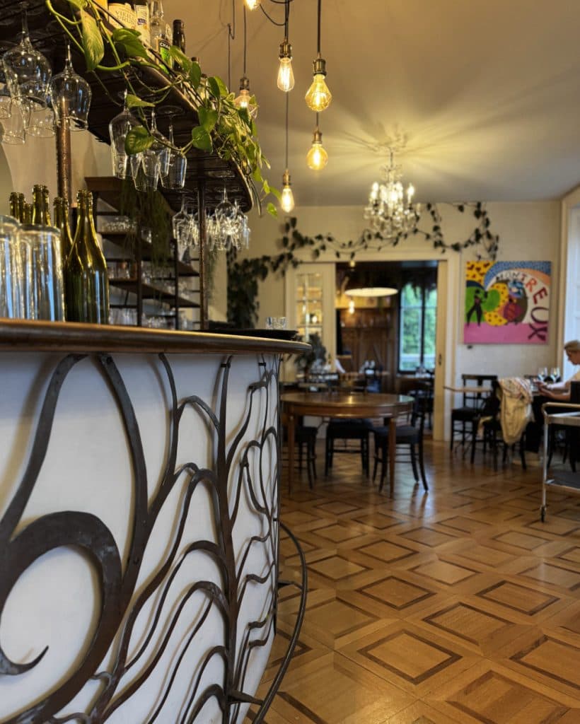 Een gezellig restaurantinterieur in Morges met houten parketvloeren, een witte bar met zwarte, wervelende metalen accenten, boven het hoofd hangende wijnglazen, groene planten, zichtbare gloeilampen en kunstwerken. Tafels en stoelen vullen de warm verlichte ruimte in het kanton Vaud.