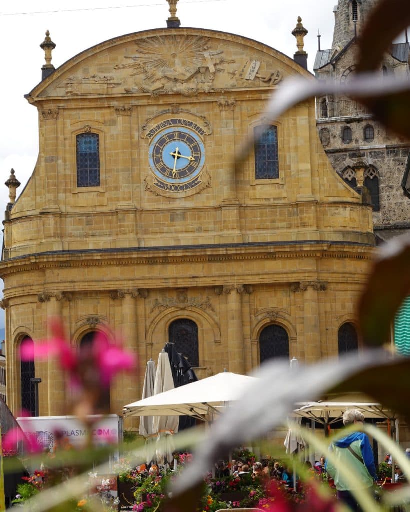 Een historisch gebouw van gele steen in Yverdon-les-Bains, in het kanton Vaud, met een sierlijke klok en een Latijnse inscriptie. Voor het gebouw staan terrastafels met parasols en kleurrijke bloemen, gedeeltelijk omlijst door wazige bladeren en roze bloesems.