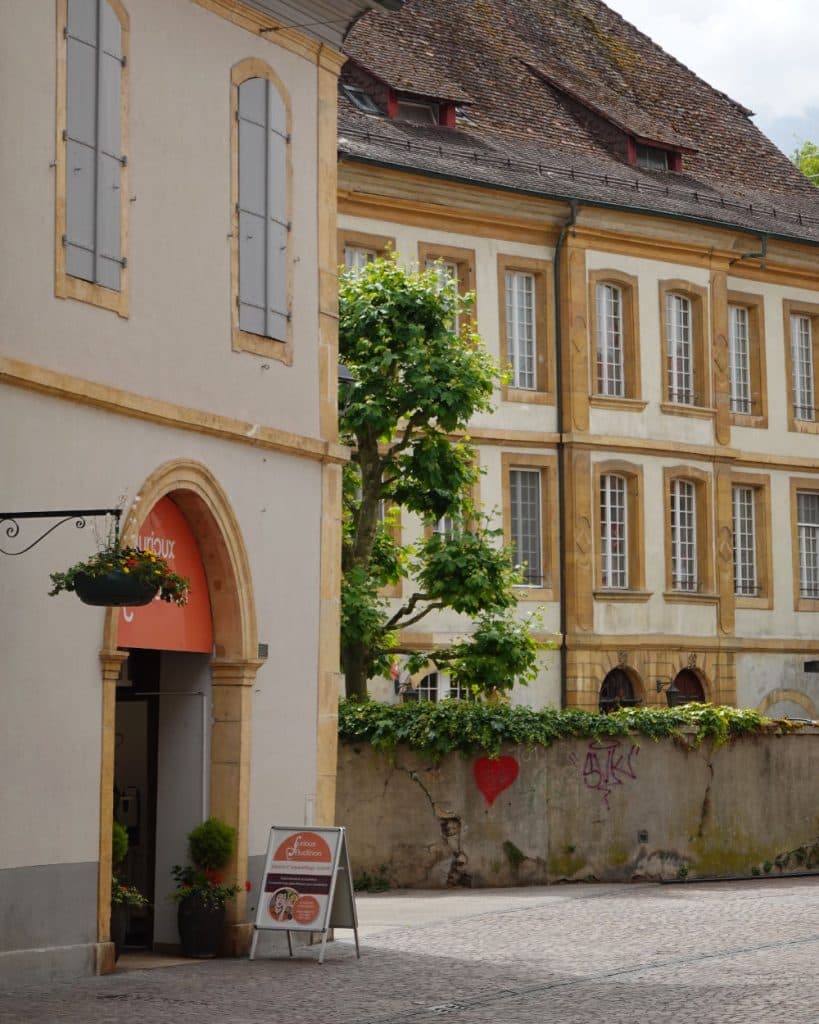 Een rustige straat met kasseien in Yverdon-les-Bains, in het kanton Vaud, met historische gele en beige gebouwen. Eén gebouw heeft hoge ramen en een boom ervoor. Links is bij een open winkeldeur een hangplant te zien, terwijl graffiti en een hart een stenen muur sieren.