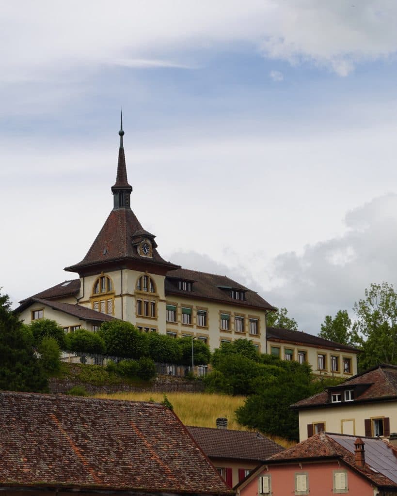 Op een met gras begroeide heuvel in Yverdon-les-Bains, in het kanton Vaud, staat een groot, historisch gebouw met een hoge, puntige klokkentoren en een steil, pannendak, omringd door bomen. Op de voorgrond, onder een bewolkte hemel, staan kleinere huizen met rode daken.