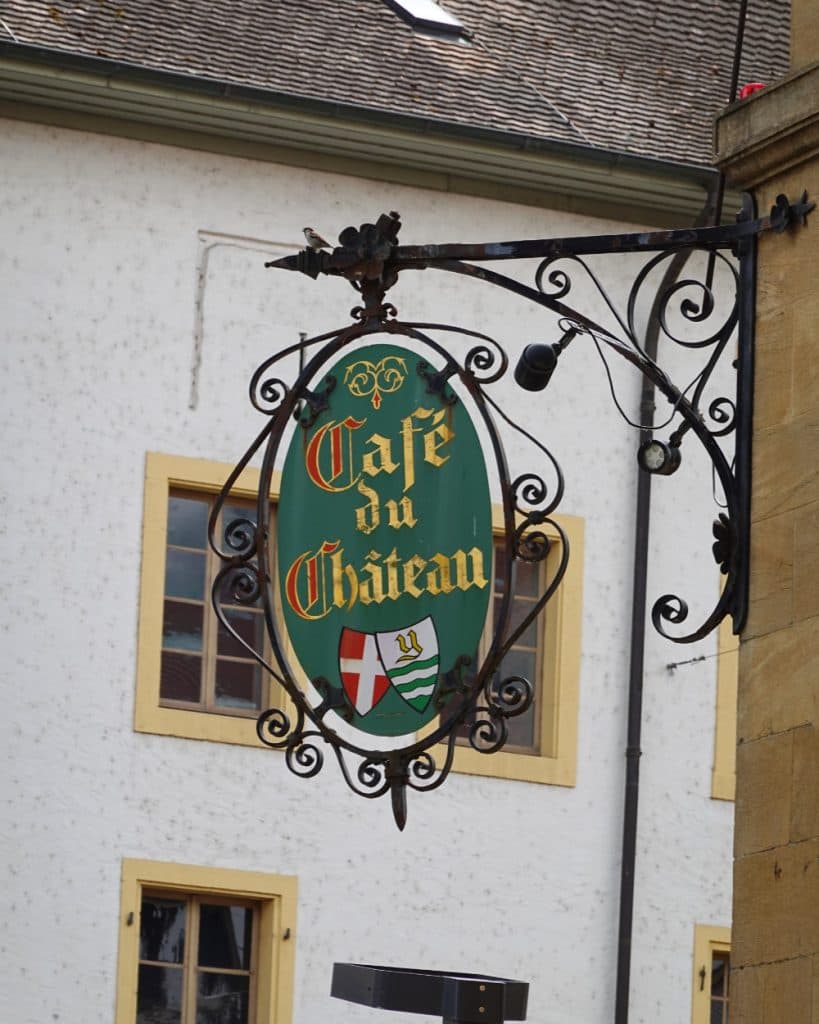 In Yverdon-les-Bains, in het kanton Vaud, hangt buiten een sierlijk metalen bord met de tekst „Café du Château” in goudkleurige letters in middeleeuwse stijl. Het groene, ovale bord is voorzien van twee heraldische schilden en is bevestigd aan een decoratieve zwarte beugel tegen een wit gebouw.