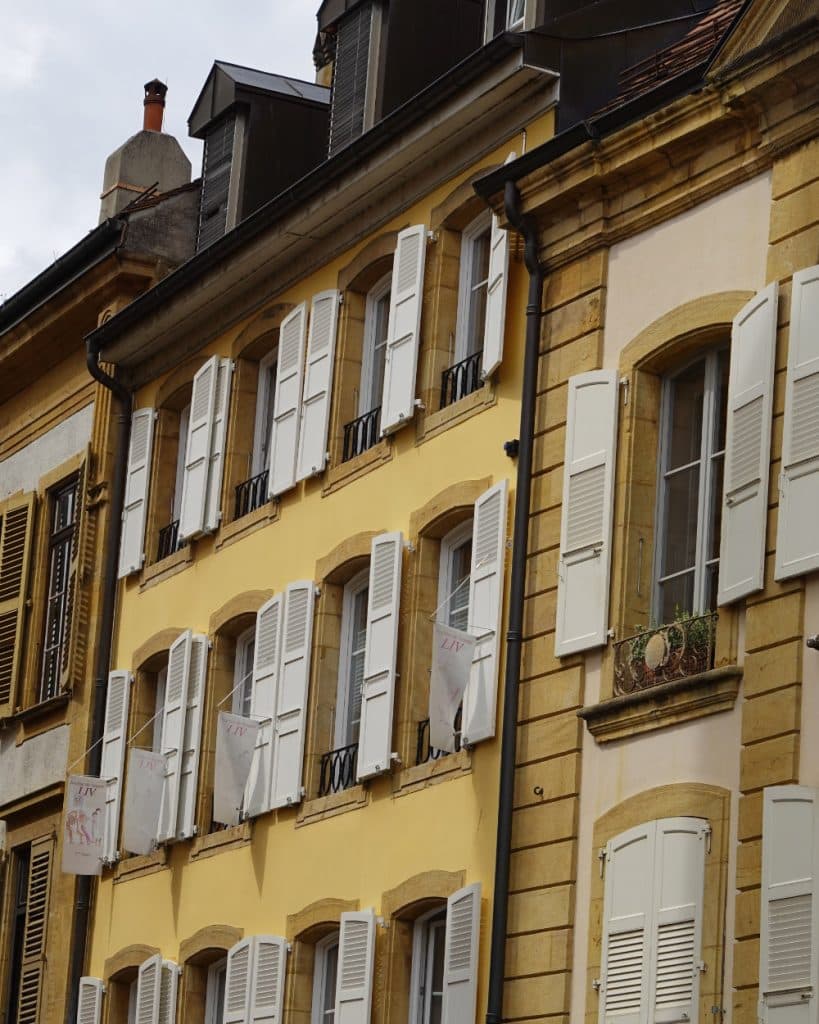 Een close-up van gele en bruine gebouwen in Europese stijl in Yverdon-les-Bains, Vaud, met witte luiken – sommige open, andere dicht. Een klein balkon met een potplant steekt af tegen de leien daken onder een gedeeltelijk bewolkte hemel.