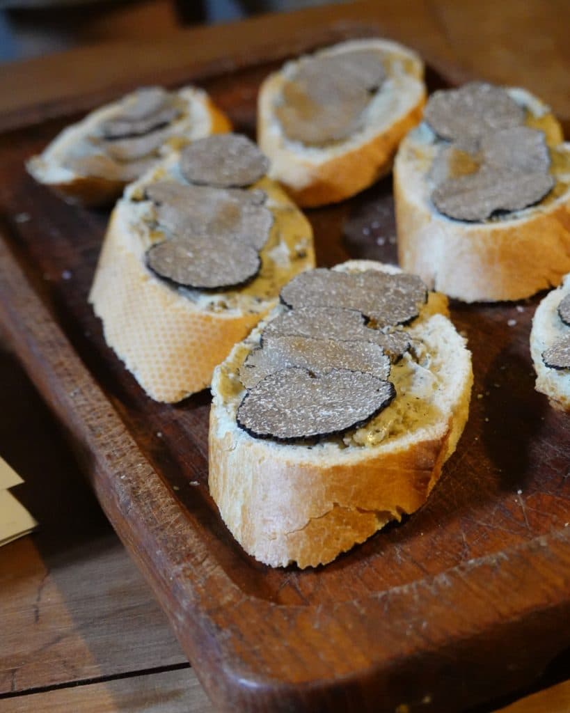 Op een aantal sneetjes stokbrood is een romige smeersel aangebracht en zijn dunne plakjes zwarte truffel gelegd; het geheel is op een houten serveerplank geschikt op een rustieke tafel in het hart van Vaud.