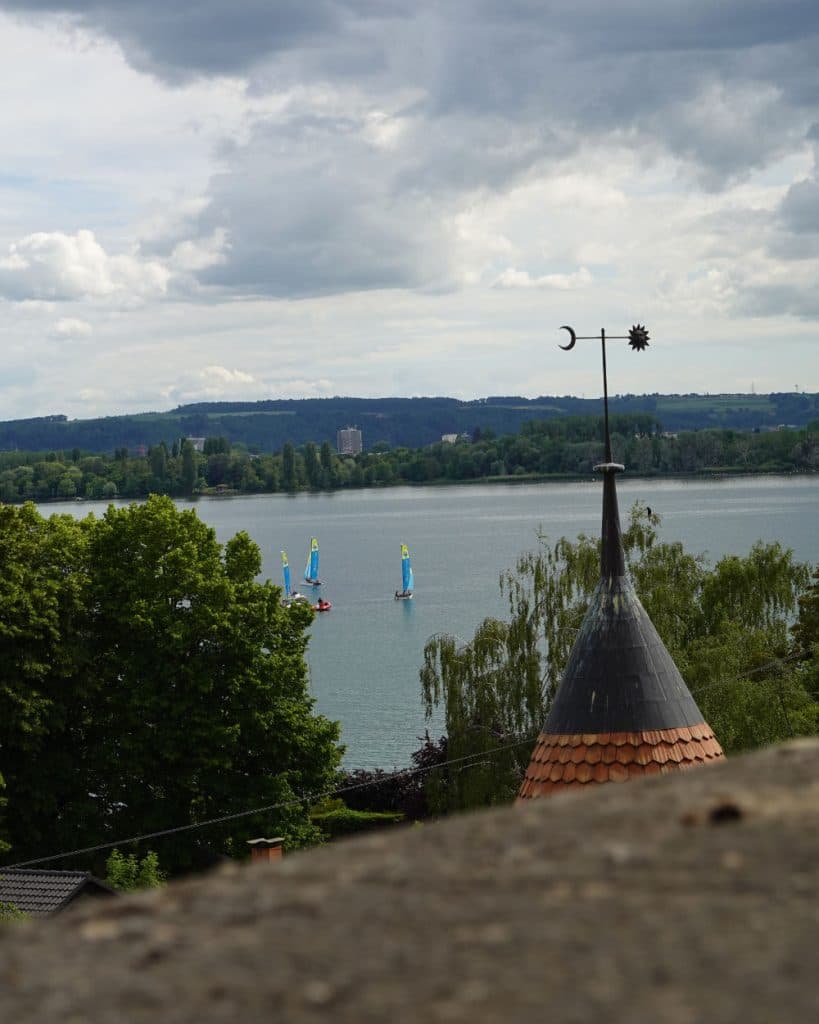 Uitzicht op een rustig meer bij Yverdon-les-Bains, met drie kleine zeilbootjes met felblauwe zeilen. Op de voorgrond steekt tussen de bomen een puntig torendak met een windwijzer uit, terwijl de heuvels en gebouwen van het kanton Vaud te zien zijn onder een bewolkte hemel.