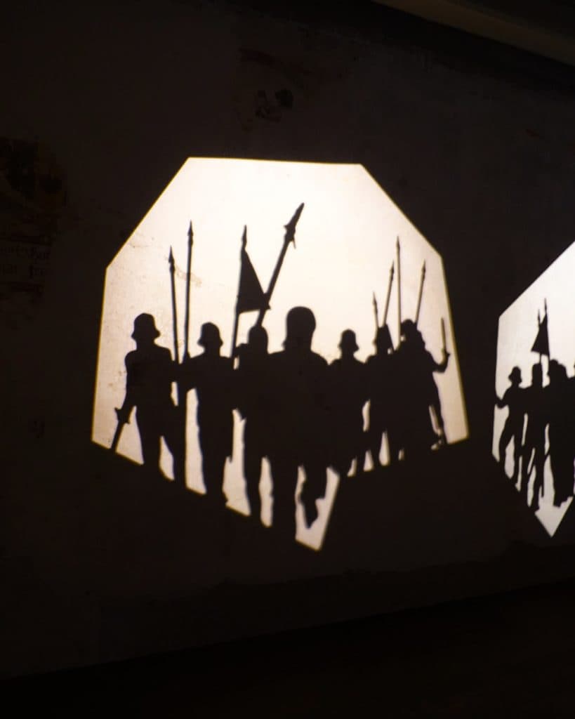 Een schaduwbeeld van soldaten met speren in de hand, die staan en voorwaarts marcheren. De figuren tekenen zich af tegen een helder achthoekig licht op een donkere muur in Yverdon-les-Bains, Vaud, waardoor een dramatisch en dynamisch schaduweffect ontstaat.
