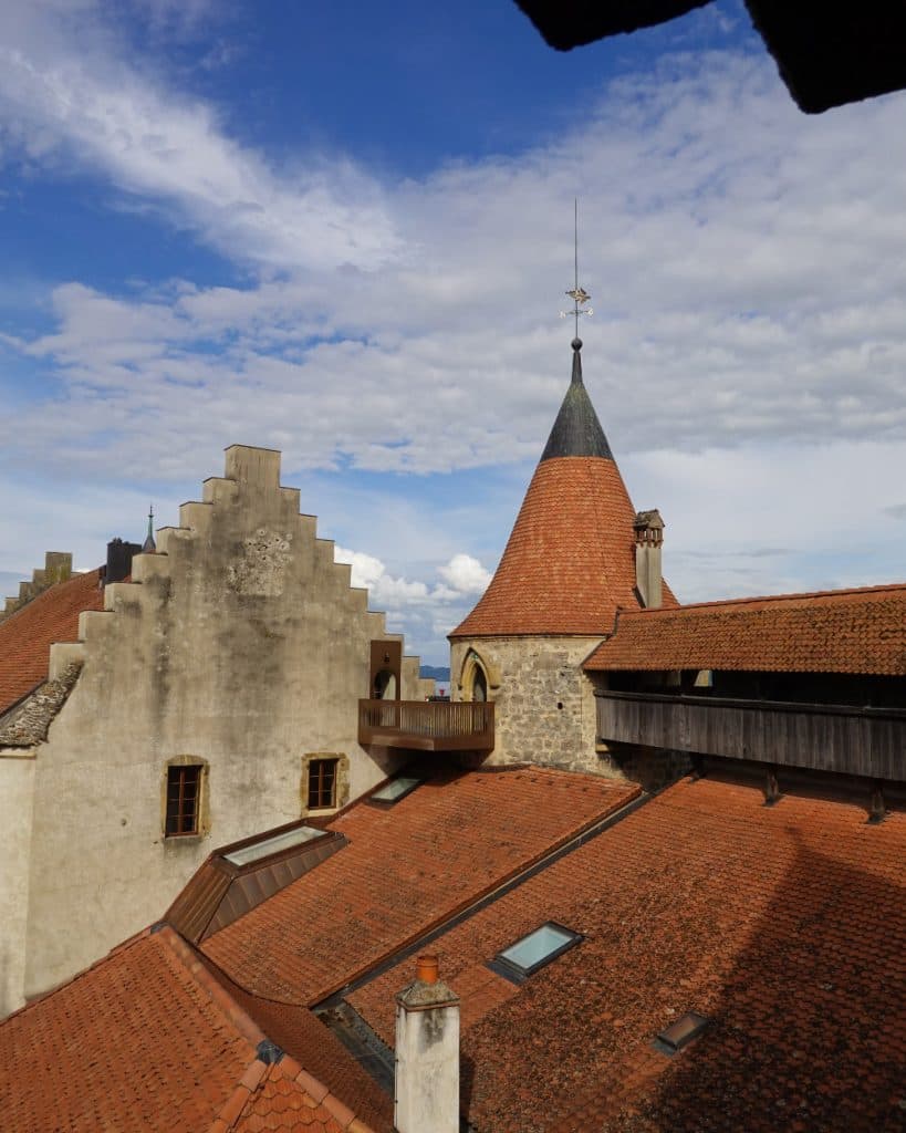 Een aanzicht van een middeleeuws stenen gebouw in Yverdon-les-Bains, in het kanton Vaud, met rode pannendaken, een kegelvormige toren en getrapte gevelwanden. Boven het gebouw is een helderblauwe lucht met hier en daar wat wolken te zien, en uit de torenmuur steekt een houten balkon uit.