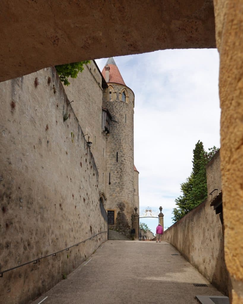 Een smal stenen pad in Yverdon-les-Bains loopt omhoog naar een middeleeuwse toren met een rood kegelvormig dak. Vlak bij de top loopt iemand in een felroze jurk, omringd door eeuwenoude bogen en de verweerde stenen muren van het kanton Vaud, met daarachter bomen en een bewolkte hemel.