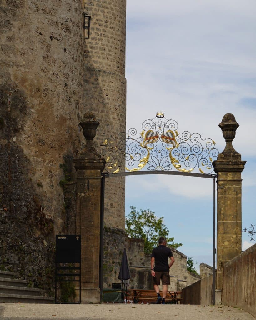 Een persoon in een korte broek en een zwart shirt loopt naar een sierlijke, met goud versierde ijzeren poort die tussen stenen pilaren staat, naast een oude stenen muur in Yverdon-les-Bains, Vaud. Achter de poort staan buitentafels, stoelen en een boom onder een gedeeltelijk bewolkte hemel.