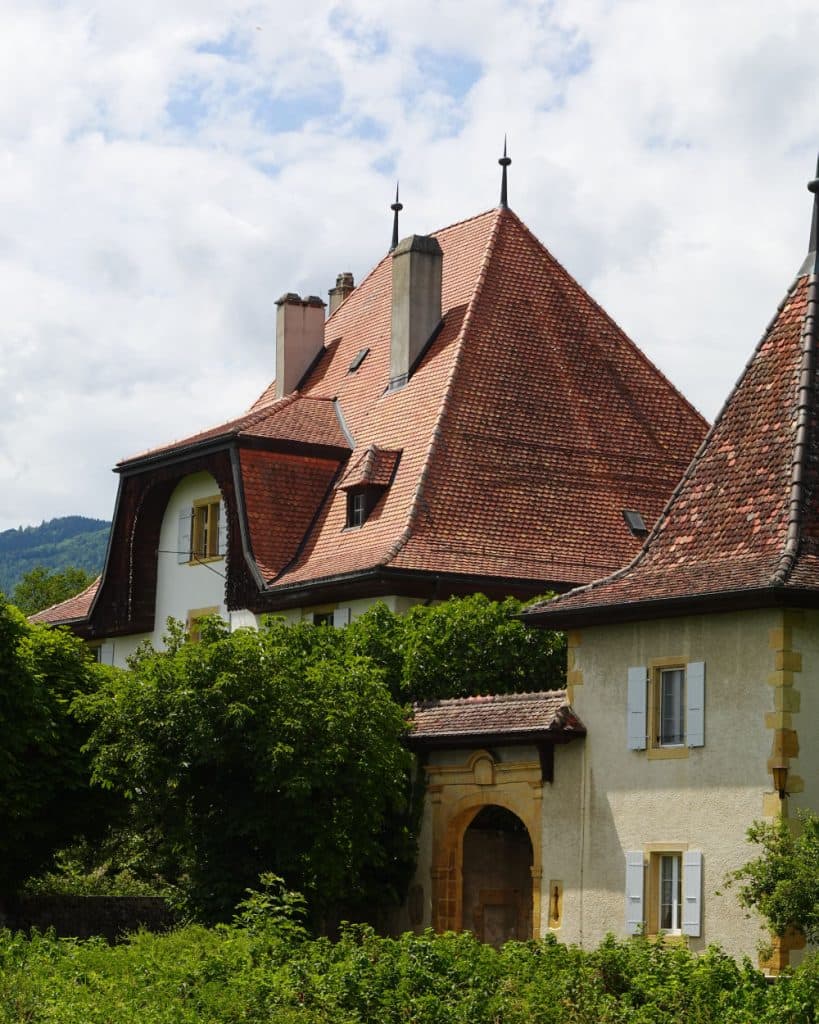 In Yverdon-les-Bains, in het kanton Vaud, staat een gebouw met steile daken met rode dakpannen, hoge schoorstenen en witte muren die gedeeltelijk door groene bomen worden bedekt. Het heeft kleine ramen en bij de ingang bevindt zich een gele stenen boog. De lucht is bewolkt en op de achtergrond zijn heuvels te zien.