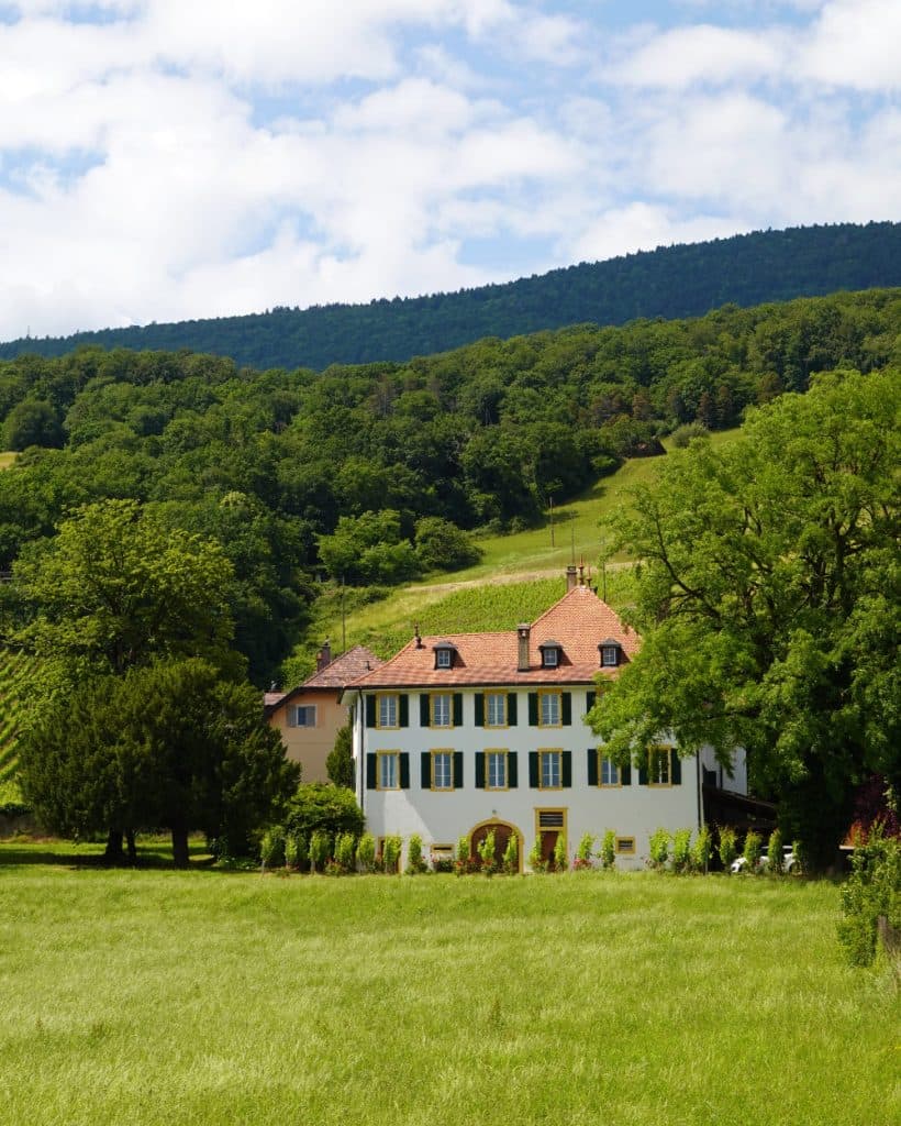Een groot wit huis met blauwe luiken en een rood pannendak staat te midden van weelderig groen, bomen en een grasveld in het rustige platteland van Vaud. Op de achtergrond rijzen beboste heuvels op onder een gedeeltelijk bewolkte hemel, wat de schilderachtige charme van Yverdon-les-Bains oproept. Het huis heeft verschillende schoorstenen en elegante gewelfde deuropeningen.