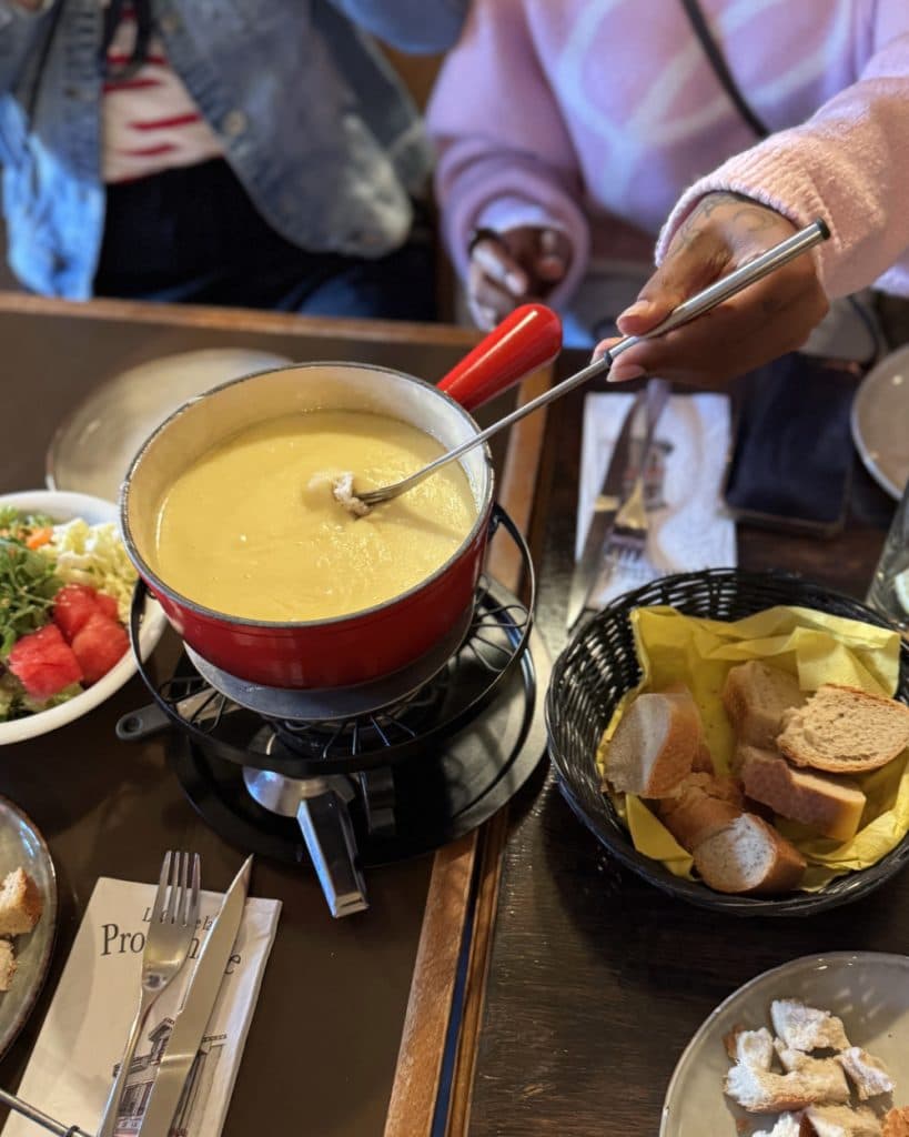 Iemand doopt brood in een rode fonduepan gevuld met gesmolten kaas, wat doet denken aan de culinaire tradities van het kanton Vaud. Op een houten tafel staan borden met salade en watermeloen, een zwarte mand met nog meer brood en bestek. Op de achtergrond zijn twee mensen gedeeltelijk te zien, die wellicht samen genieten van deze gezellige maaltijd in Yverdon-les-Bains.