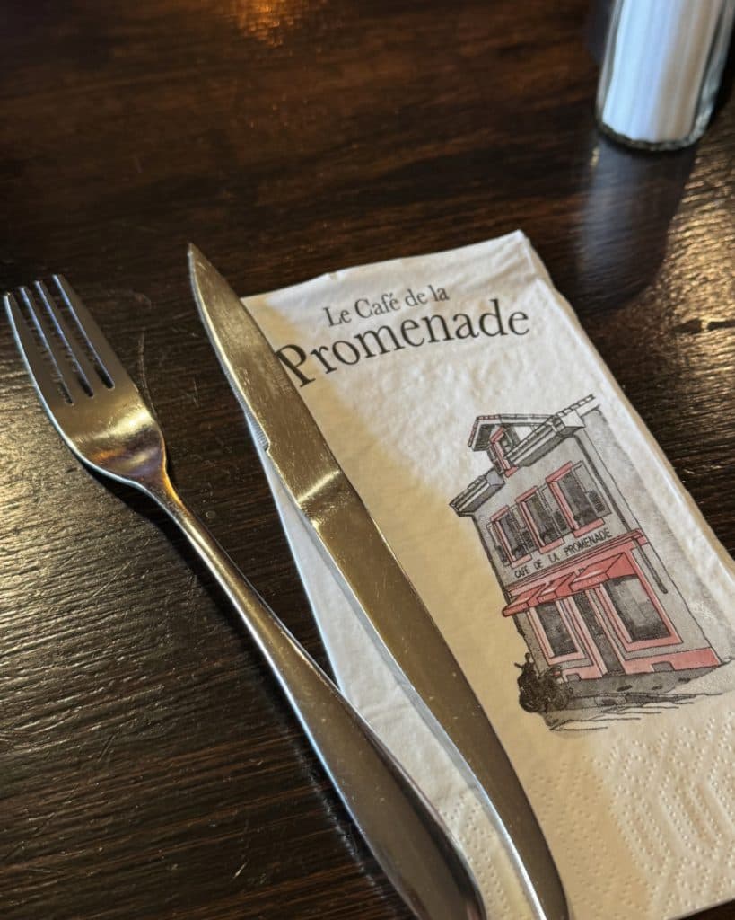 Op een donkere houten tafel liggen een vork en een mes op een servet met de tekst „Le Café de la Promenade“ en een afbeelding van een roze en grijs cafégebouw, dat doet denken aan de charmante straatjes van Yverdon-les-Bains in Vaud. In de rechterbovenhoek is een deel van een cilindervormige bak te zien.