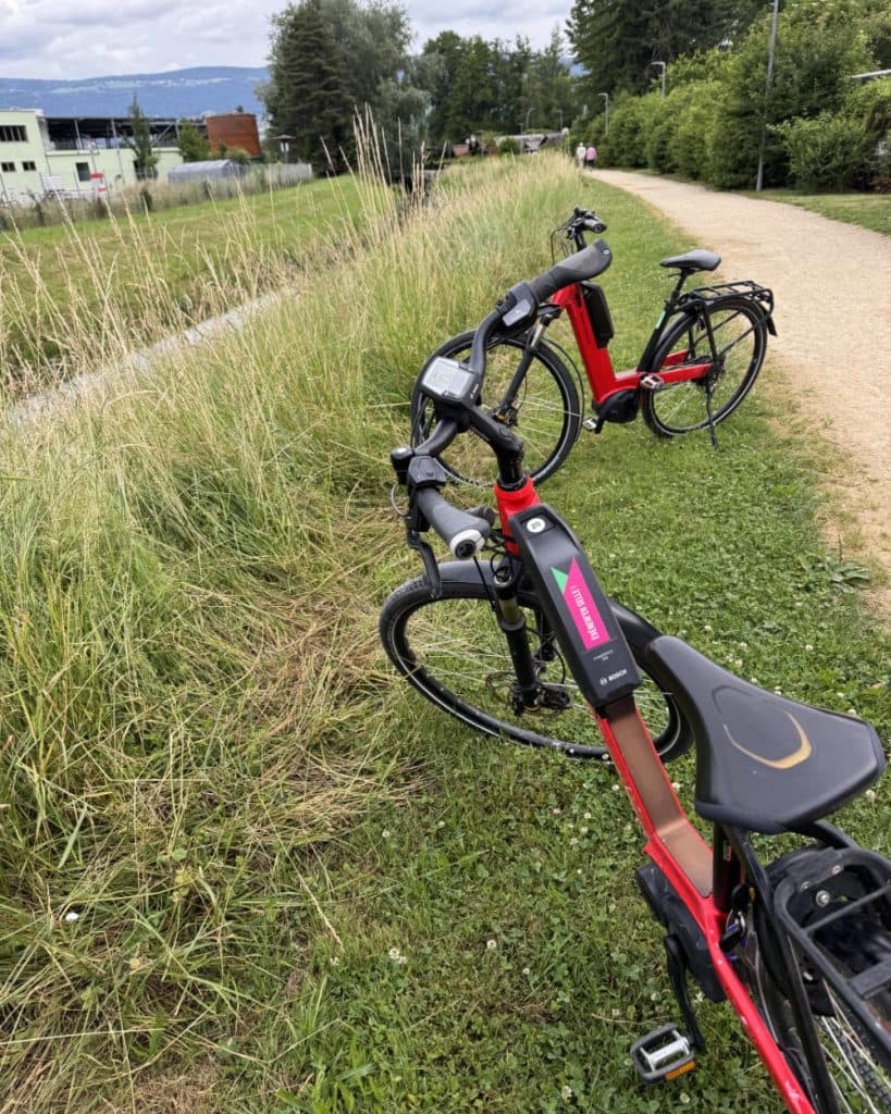 Er staan twee rode elektrische fietsen op het gras naast een onverhard pad dat omzoomd wordt door hoog gras. Op de achtergrond zijn bomen, gebouwen en een gedeeltelijk bewolkte hemel te zien. Het tafereel doet denken aan een rustig park of een landelijk gebied in het kanton Vaud, in de buurt van Yverdon-les-Bains.