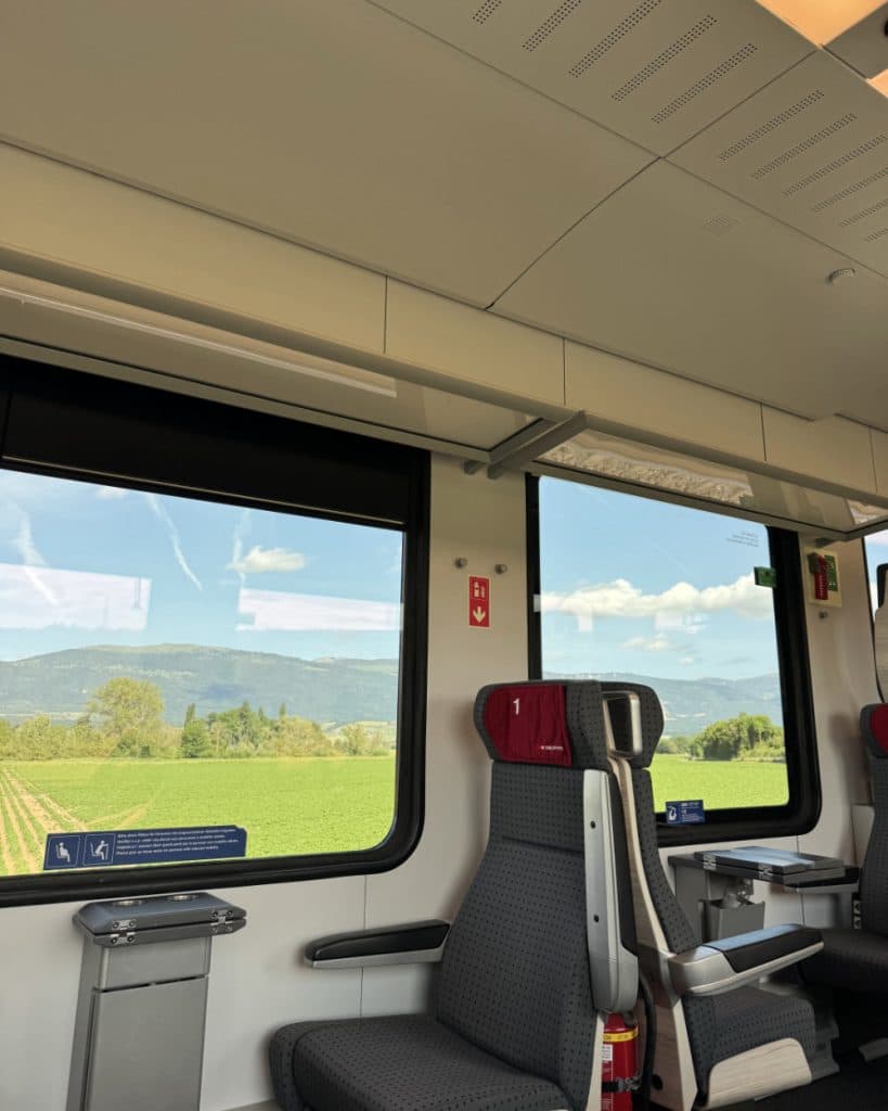 Het interieur van de trein met grijze stoelen en rode hoofdsteunen, grote ramen die uitzicht bieden op groene velden, bomen en heuvels in de verte onder een blauwe lucht met wolken; naast de stoelen zijn noodborden en een klein tafeltje te zien — een typisch tafereel op een traject dat door het kanton Vaud loopt, in de buurt van Yverdon-les-Bains.