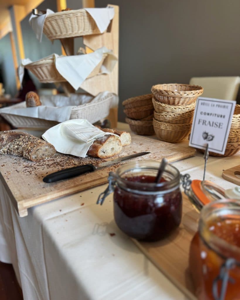 Gesneden meergranenbagette op een houten plank met een mes, een pot aardbeienjam, sinaasappelmarmelade, rieten mandjes en een broodrek op een tafel. Op een klein bordje staat „Confiture Fraise“. De uitnodigende setting lijkt een ontbijtbuffet te zijn in Yverdon-les-Bains, Vaud.