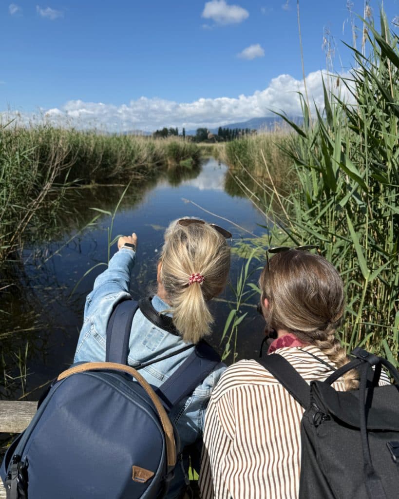 Twee mensen met rugzakken staan bij een houten reling in de buurt van Yverdon-les-Bains in Vaud, met uitzicht op een rustig stromend beekje dat omringd wordt door hoog riet. De ene wijst naar voren, de andere kijkt in dezelfde richting. De lucht is blauw met hier en daar wat wolken en op de achtergrond zijn bergen in de verte te zien.