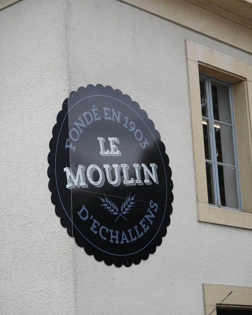 Op een ronde zwarte bord op een beige muur staat in witte letters „Fondé en 1903 Le Moulin d'Echallens”, met onder de naam een tarwesymbool dat verwijst naar de rijke geschiedenis van Vaud. Naast het bord is een raam met een beige kozijn te zien.