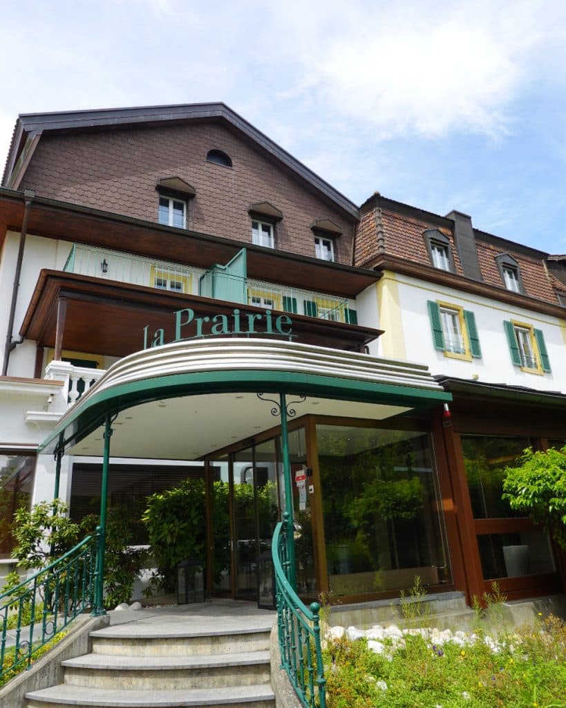Een hotelingang met groene leuningen, glazen deuren en een bord met het opschrift „la Prairie“ heet gasten welkom in Yverdon-les-Bains. Het gebouw heeft bruine dakspanen op de bovenste verdieping, witte muren, groene luiken en wat groen rondom de trap – dit alles tegen de achtergrond van de gedeeltelijk bewolkte hemel boven Vaud.