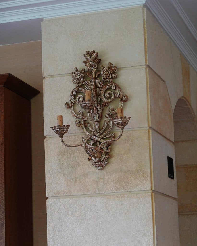 Een sierlijke, goudkleurige wandlamp met gedetailleerde bloem- en bladmotieven is bevestigd aan een beige stenen muur in een verfijnde woning in Yverdon-les-Bains. De wandlamp is voorzien van twee kaarsvormige lampen en hangt tussen een houten kast en een gewelfde deuropening, wat de elegante charme oproept die vaak in interieurs in het kanton Vaud te vinden is.