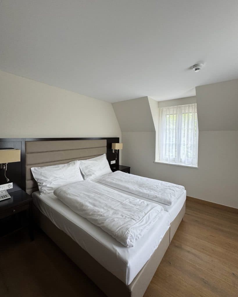 In een lichte kamer met houten vloeren staat een keurig opgemaakt tweepersoonsbed met wit beddengoed, een donker hoofdeinde, twee nachtkastjes met lampjes en een raam met vitrages die het daglicht binnenlaten – een rustgevende omgeving die doet denken aan de gastvrije interieurs die je vaak aantreft in Yverdon-les-Bains, in het kanton Vaud.