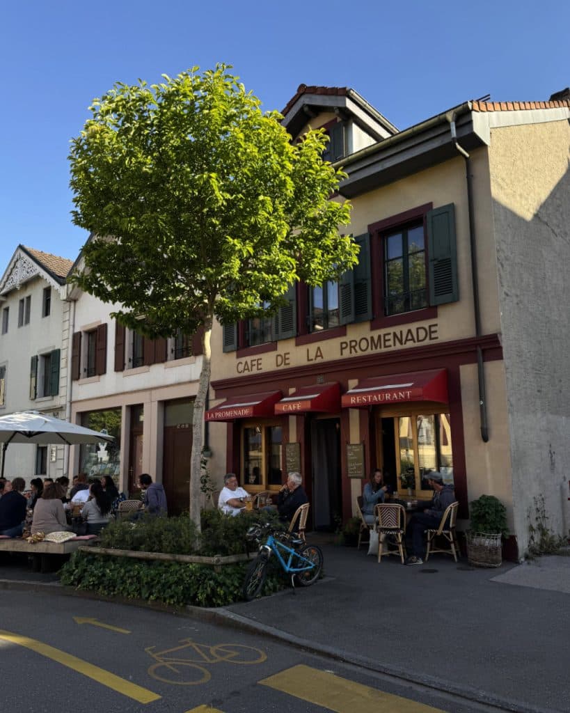Op een zonnige dag in Yverdon-les-Bains, in het kanton Vaud, zitten mensen buiten bij Café de la Promenade, een gebouw van twee verdiepingen met rode luifels en groene luiken. Een boom zorgt voor schaduw op het trottoir en vlakbij staat een fiets geparkeerd. Op de straat ervoor is een geel fietsstrookteken geschilderd.