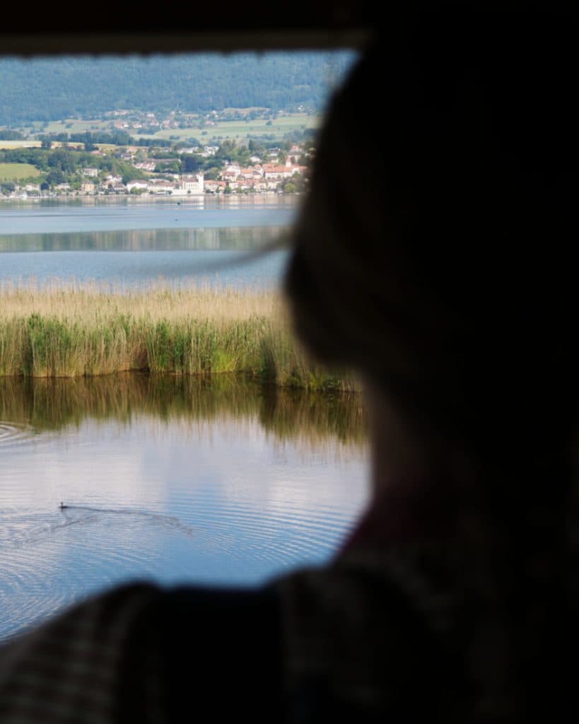 Een figuur in de schaduw kijkt vanuit een raam uit over een rustig meer, omzoomd door hoog riet. Een klein vogeltje zwemt op het water. Aan de overkant van het meer, vlakbij Yverdon-les-Bains in het kanton Vaud, ligt een afgelegen dorpje onderaan groene heuvels, onder een heldere hemel.