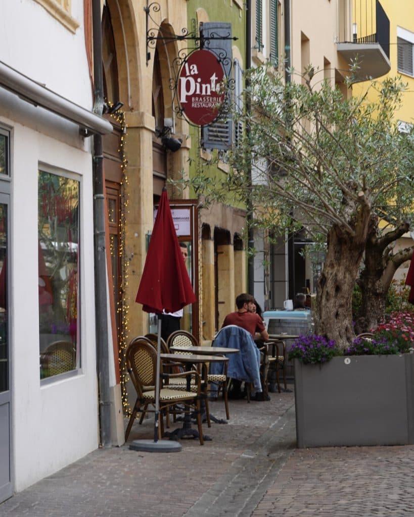 Een smalle straat met kasseien in Yverdon-les-Bains, in het kanton Vaud, staat vol met cafétafels en -stoelen, waarvan sommige in de schaduw van rode parasols staan. Een stel zit samen aan een tafel, terwijl lichtslingers een gebouw versieren waarop het uithangbord van restaurant „La Pint“ hangt. Potplanten en een grote boom geven de wandelstrook extra charme.