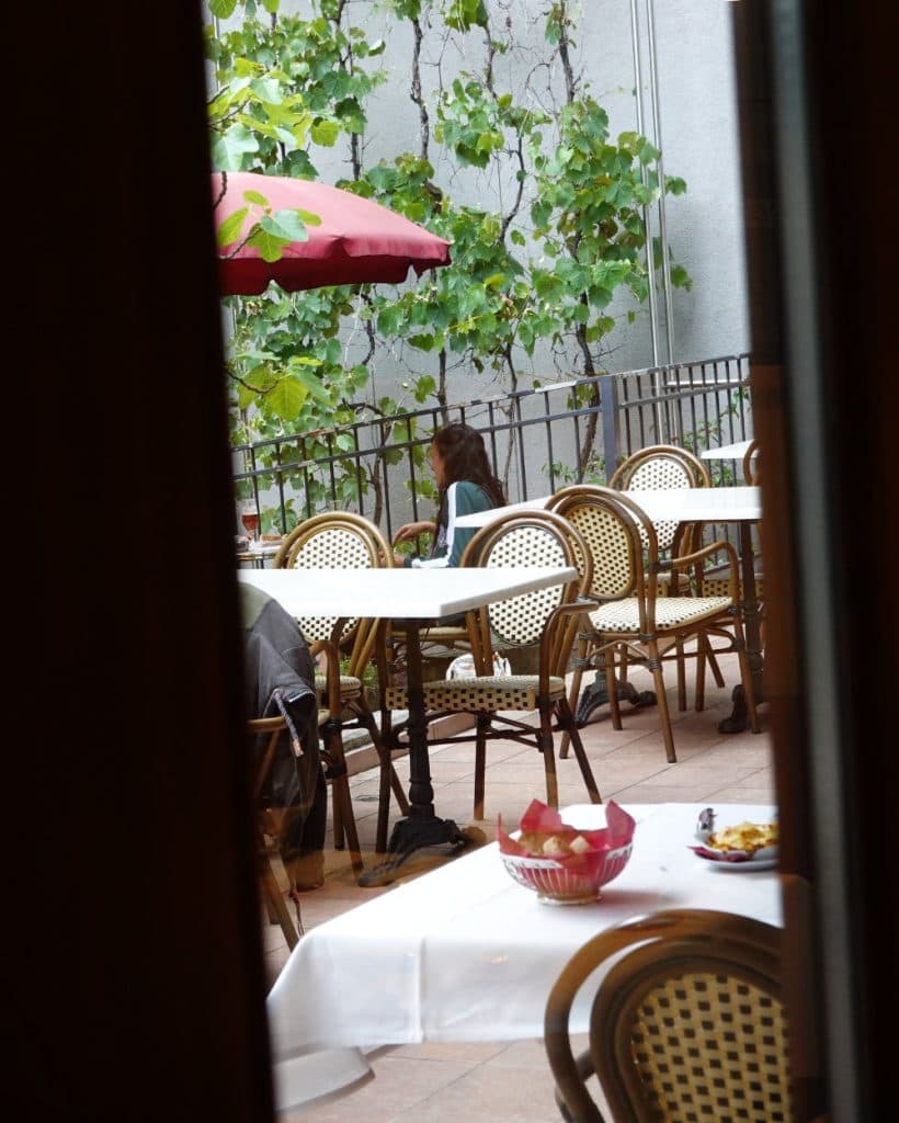 Op het terras van een café in Yverdon-les-Bains staan lege rotanstoelen en witte tafels. Een persoon met lang haar zit daar alleen, met de rug naar de kijker, omringd door het rustige groen en een opvallende rode parasol buiten. Op de voorgrond staan een kom en een bord met eten op een tafel, gedeeltelijk zichtbaar door een deuropening – een uitnodigend tafereel in het hart van Vaud.
