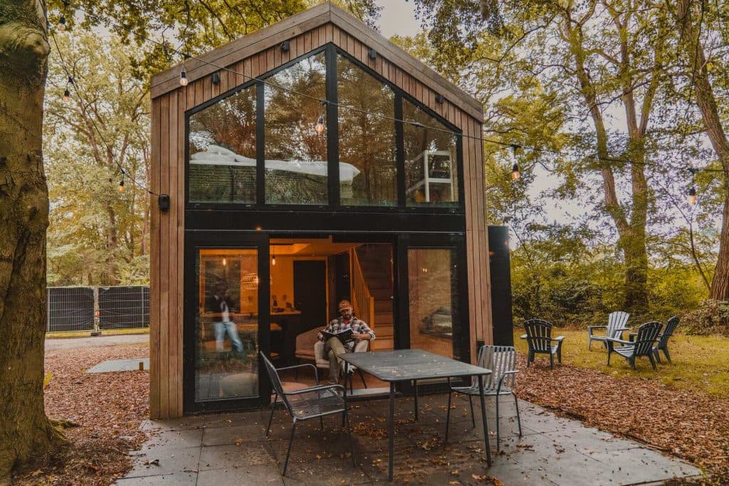 Iemand zit aan een tafel op het terras van een moderne blokhut met twee verdiepingen en grote ramen, omringd door bomen en tuinstoelen, en geniet van de rust van een overnachting in het hart van de Brabantse natuur.