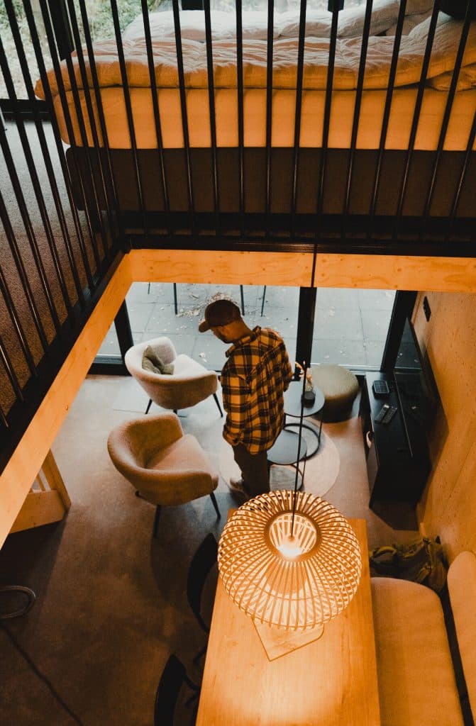 Een persoon in een geel geruit overhemd staat in een moderne loft met een bed op een tussenverdieping, ronde stoelen, een tafel en een brandende hanglamp, waardoor de ruimte een vleugje van de eigentijdse stijl van Breda krijgt.