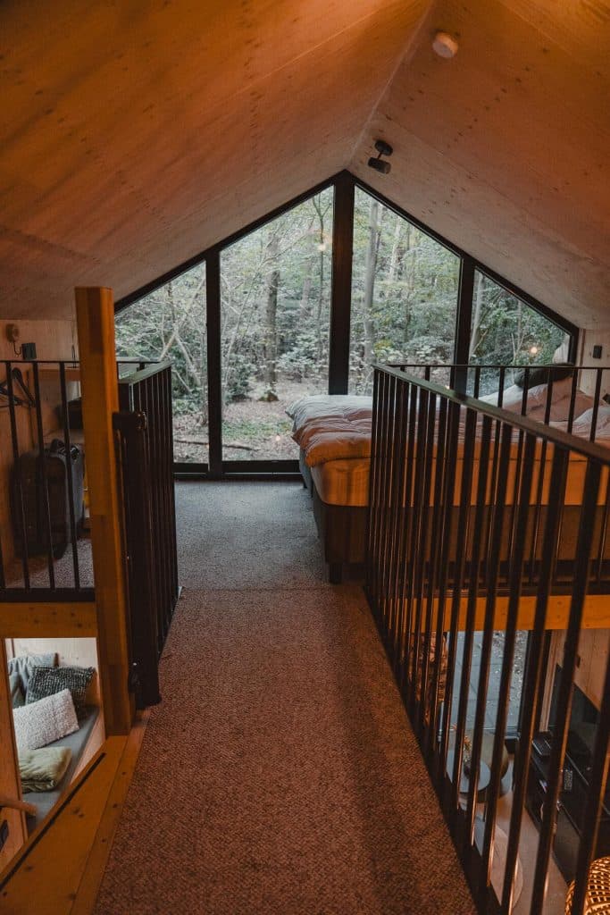 Loftslaapkamer met een groot driehoekig raam dat uitkijkt op de rustige Brabantse natuur, met tapijt op de vloer, een houten plafond en aan beide zijden zwarte metalen leuningen. Het bed staat tegen het raam aan en biedt een unieke overnachtingservaring in de buurt van Breda.