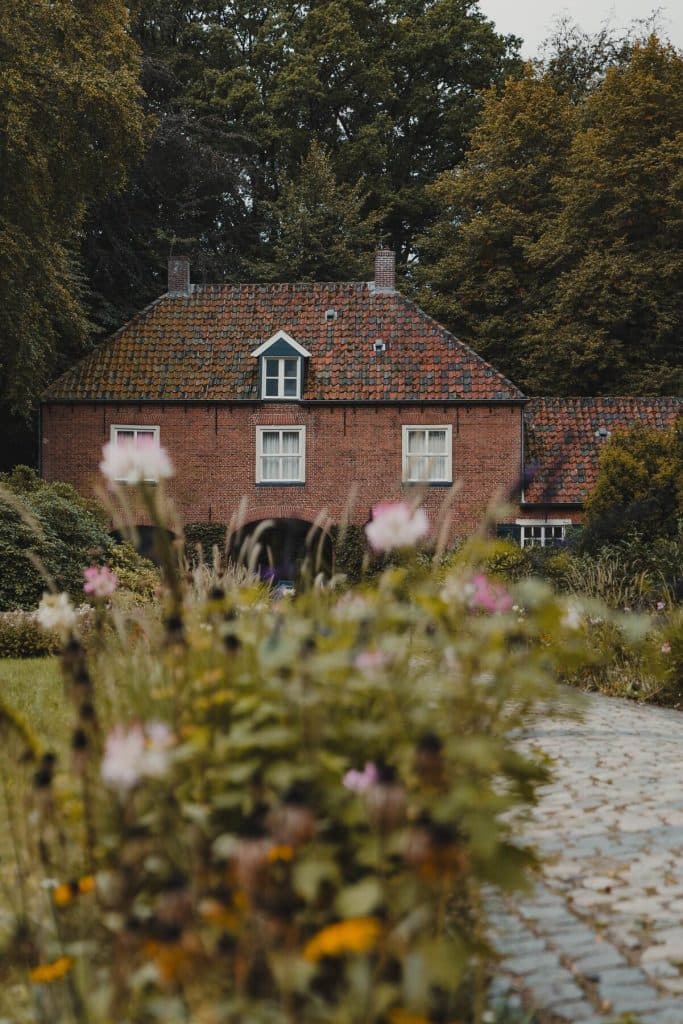 Achter een tuin met wilde bloemen en een stenen pad staat een huis van rode baksteen met een rood pannendak, omringd door hoge groene bomen – een idyllische plek om te overnachten en te genieten van de schoonheid van de Brabantse natuur in de buurt van Breda.