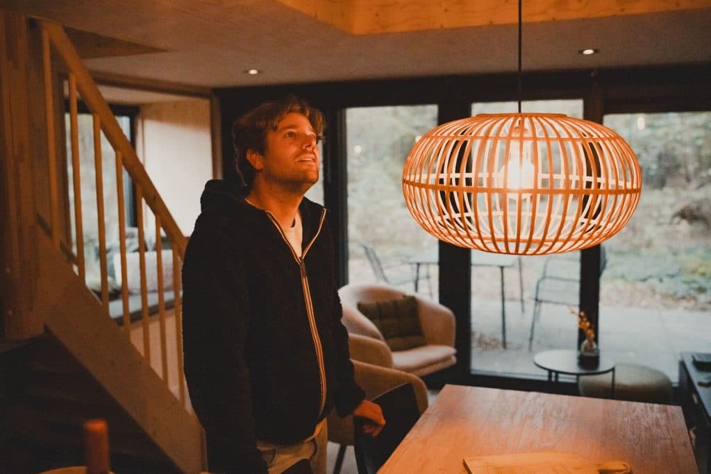 Een man in een zwarte hoodie kijkt omhoog naar een brandende hanglamp in een moderne, warm verlichte woonkamer met grote ramen en houten accenten – een rustgevende ruimte die de serene sfeer van de Brabantse natuur oproept.