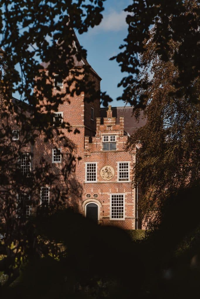 Een roodbakstenen gebouw met grote ramen en sierlijk metselwerk, gedeeltelijk omlijst door boomtakken en bladeren op de voorgrond, dat een charmant kijkje biedt in de Brabantse natuur – een ideale plek om te overnachten en te eten of om te genieten van een glas nonnenwijn.