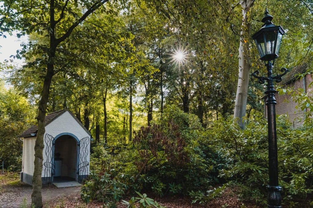 Tussen de dichte bomen en struiken, vlakbij een zwarte lantaarnpaal, staat een klein wit gebouw met een gewelfde ingang, omgeven door de rustige Brabantse natuur; het zonlicht schijnt door de takken.