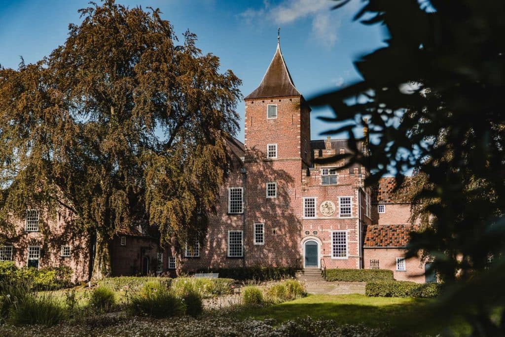 Een historisch bakstenen gebouw met een centrale toren, een grote boogdeur en talrijke ramen, omgeven door bomen en tuinen onder een blauwe hemel – een unieke plek om in Breda te overnachten en te genieten van de schoonheid van de Brabantse natuur.