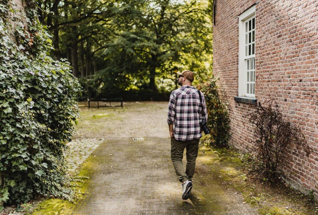 Een man in een geruit overhemd en met een pet loopt over een pad naast een bakstenen gebouw, omringd door bomen en groen, terwijl hij geniet van de rustige Brabantse natuur.