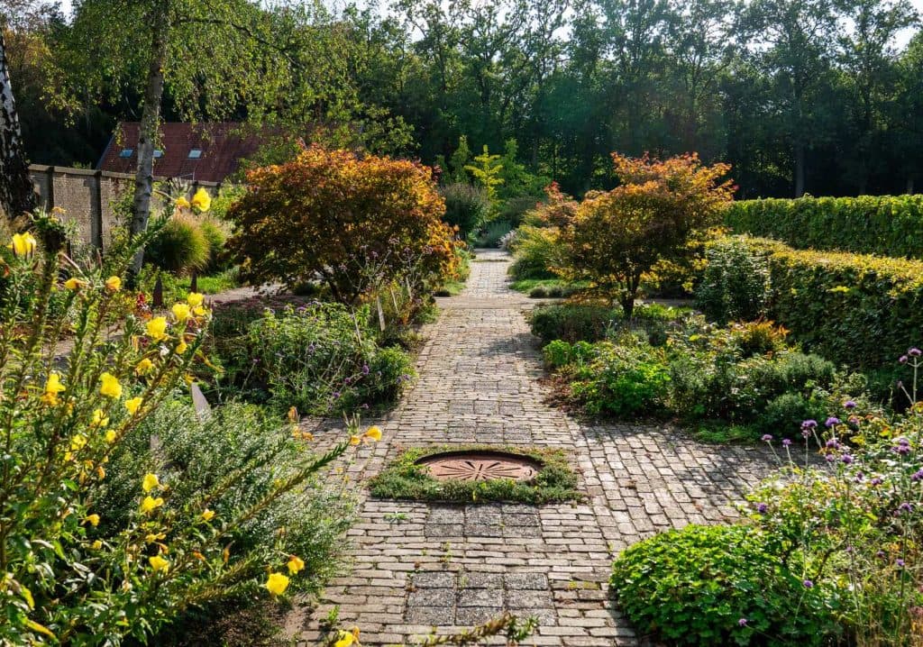 Een met bakstenen geplaveid pad slingert door een goed onderhouden tuin met diverse struiken, bloemen en bomen in het heldere daglicht, en biedt een rustige plek om te overnachten te midden van de schoonheid van de Brabantse natuur.
