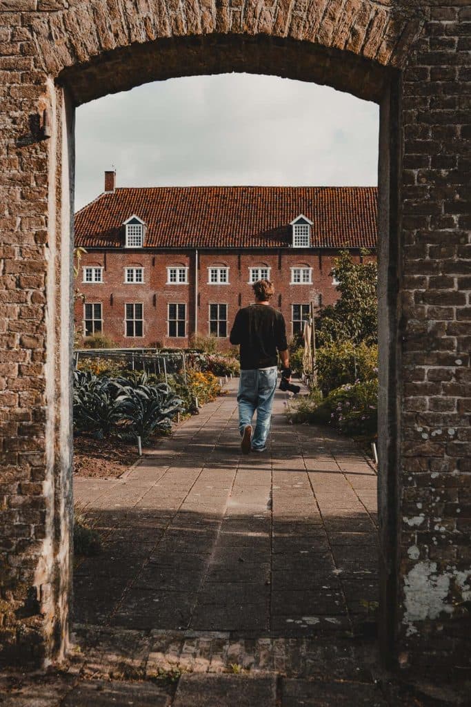 Een persoon in een spijkerbroek en een donker jasje loopt over een tuinpad naar een groot bakstenen gebouw met veel ramen en een rood pannendak, omlijst door een stenen boog – een rustig tafereel dat ‘het goede leven’ in Breda perfect weergeeft.