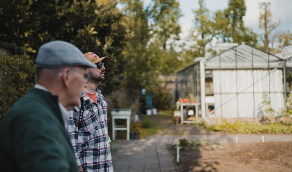 Twee mannen staan buiten bij een tuin met op de achtergrond een glazen kas, omringd door bomen en planten, en genieten van het goede leven in deze rustige omgeving – misschien proosten ze zelfs met een glas nonnenwijn terwijl ze de sfeer van Bruisend Breda in zich opnemen.