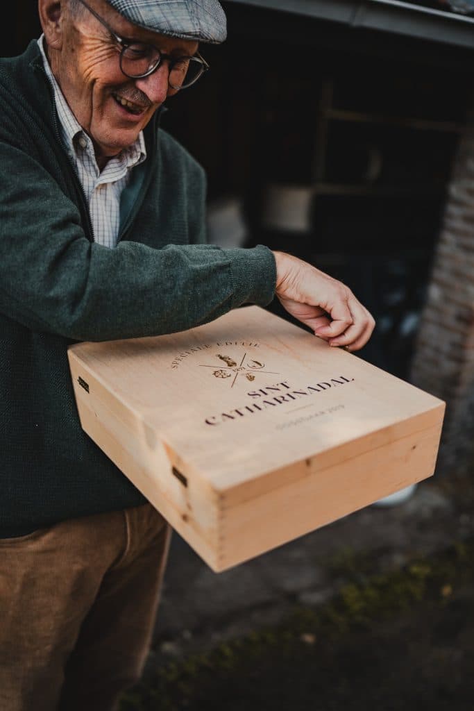 Een oudere man met een bril en een pet houdt buiten een houten kistje vast met het opschrift ‘Catherine Dal’; hij glimlacht terwijl hij het opent en geniet duidelijk van het goede leven – misschien wel klaar om een fles nonnenwijn uit het hart van Bruisend Breda te delen.