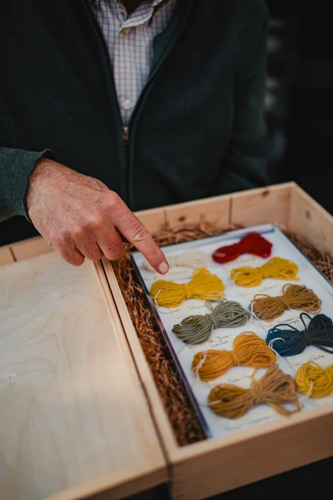 Iemand wijst naar een houten kistje met gelabelde bundeltjes gekleurd garen, keurig gerangschikt op een vel papier – een uitnodiging om ‘het goede leven’ te ervaren door middel van creativiteit en aandachtig handwerk.