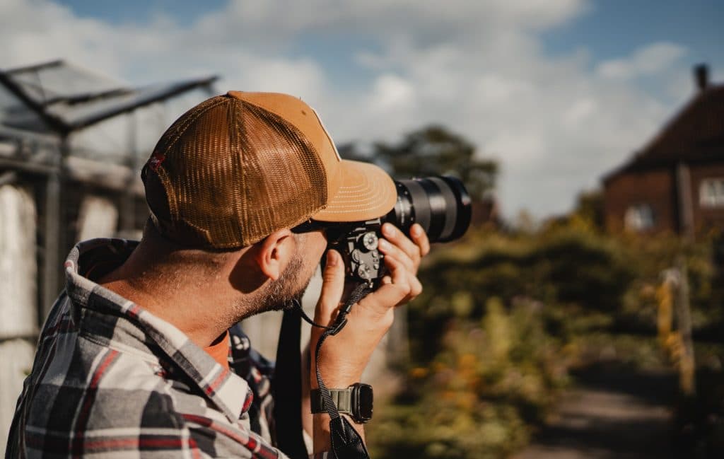 Iemand in een geruit overhemd en een bruine pet houdt een camera voor zijn gezicht en maakt op een zonnige dag in Breda buiten foto’s – om momenten vast te leggen die ‘het goede leven’ vieren.